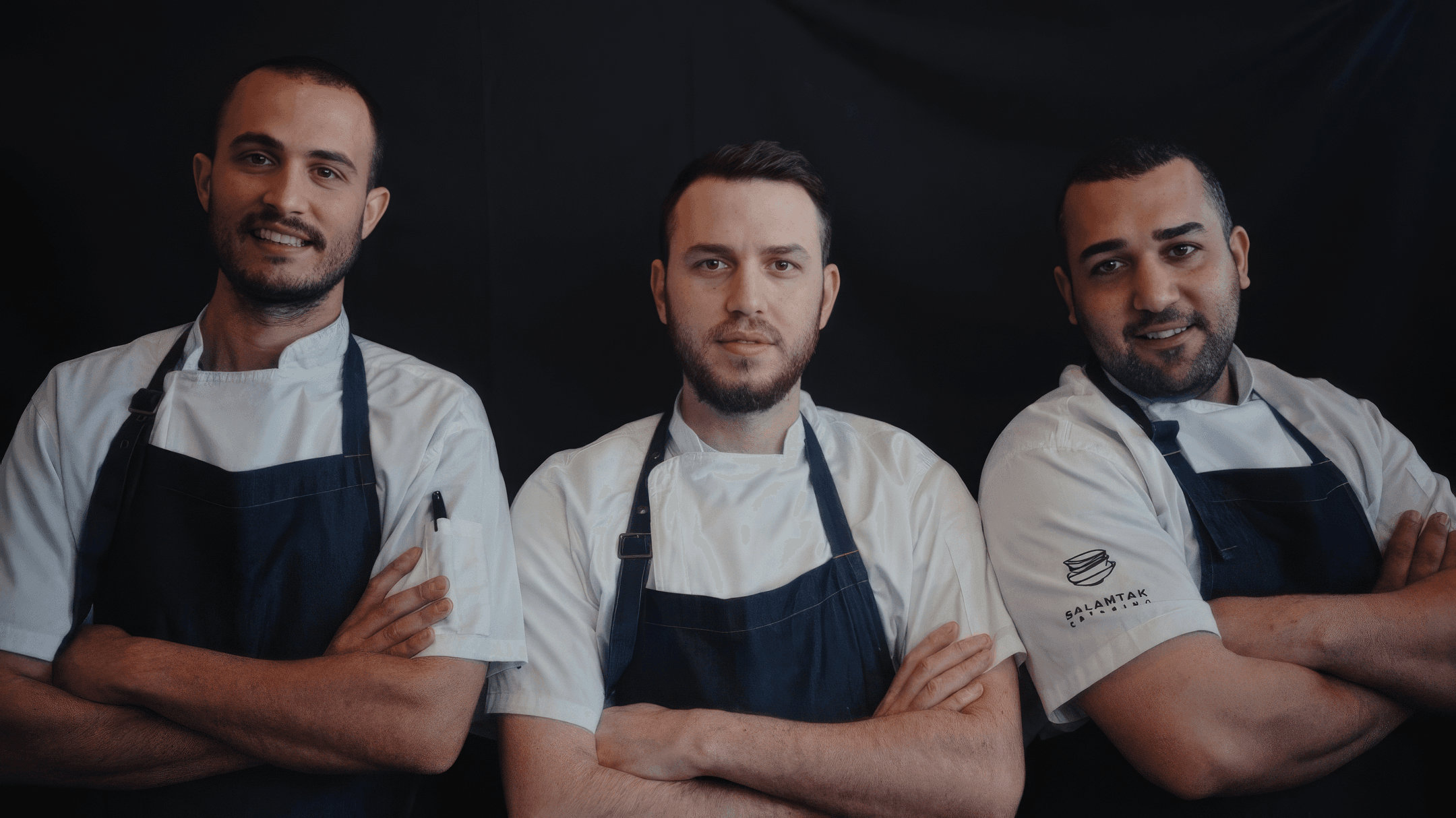 Three members of Corridor 18’s founding team stand shoulder to shoulder in white chef coats and dark aprons, arms crossed, against a black studio backdrop.